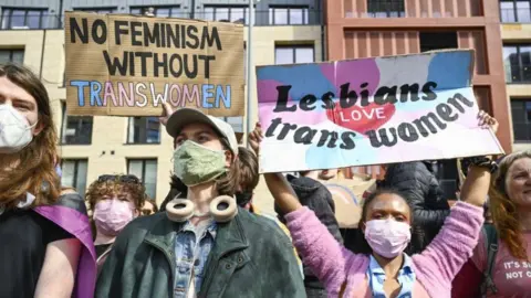 A group of protesters hold up placards. One reads "No feminism without trans women". Another reads "lesbians love trans women" and is being held up by a woman wearing a pink cardigan and a pink face mask. 