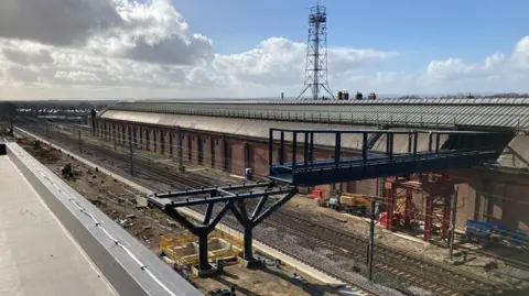 A black footbridge is being installed in sections over a single railway line at Darlington Station. Behind the footbridge is the long redbrick building which makes up part of the original station building. 