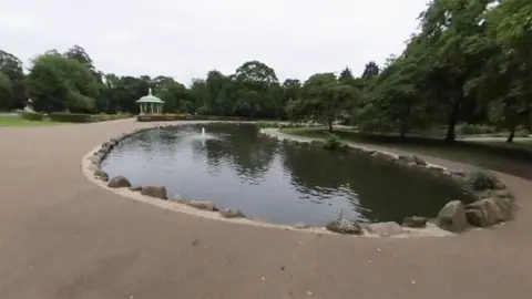 View of Pearson Park pond. It is curved and surrounded by pathways with a bandstand in the background.