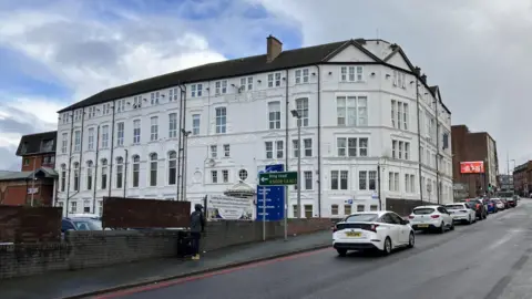 A white-painted five-storey building situated on a hill with a road running alongside it. There are cars queuing at a set of traffic lights and a person wearing a grey hoodie on the pavement. There is a road sign partially obscuring a blue totem which carries the hotel's branding and other information.