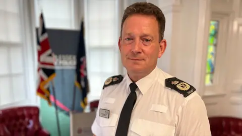 Stuart Woodward Ben-Julian Harrington stands in a white shirt with black epaulettes and a black tie. He is standing in front of a lectern with an Essex Police banner in the background.