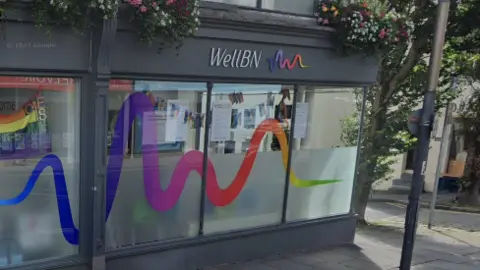 The outside of WellBN in Brighton. It is a grey-painted building with half-frosted windows and a rainbow wiggly line - its logo - on the sign. 