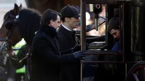 Getty Images Benedict Cumberbatch dressed as Sherlock Holmes rides in a carriage, surrounded by two other men, both in black