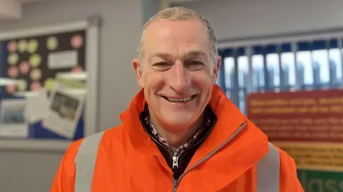A man with very little grey hair is smiling at the camera. He is wearing an orange Hi Viz coat and a black hoody and checkered shirt. 