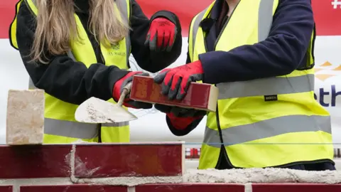 PA Media Two pairs of hands holding a brick whilst learning how to lay bricks. The people are wearing high vis jackets and red gloves.