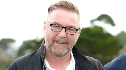 Hollywood director Michael Caton Jones - a fair-haired man with a beard and glasses stands against a leafy background, wearing a black collared jacket.