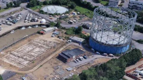 An aerial view of a leisure park. There are trees, parked cars and boats, as well as a large blue gasometer.