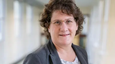 The Queen Elizabeth Hospital A woman with short curling dark hair wearing wire framed glasses and a dark grey jacket, looking towards the camera and slightly smiling