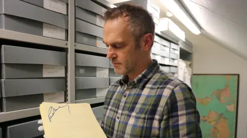Roald Dahl Museum and Story Centre A man with short brown hair and wearing a checked shirt holding a manuscript in his hands. He is wearing white gloves. There are boxes stacked on shelves behind him.