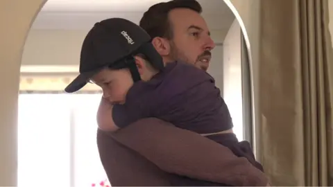 A dad holding his son - wearing a hat - over his shoulder