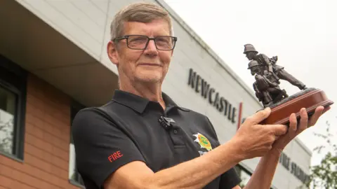 Kelvin Knapper wearing a black Staffordshire Fire and Rescue Service shirt holds up his award - a trophy featuring two miniature firefighters.