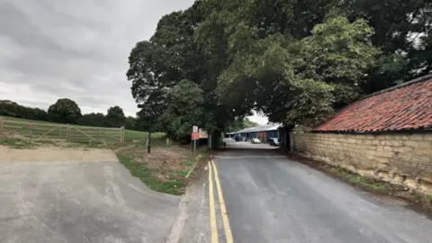 A quiet rural street with fields on the left and brick buildings on the right. Large green trees grow over the path. In the distance are some warehouse buildings