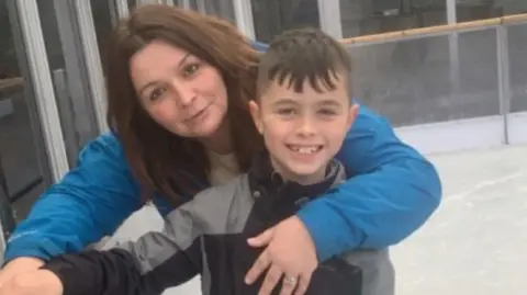 Family Noah and his mother are standing outside, possibly on an ice rink, and resting near a railing. Louise has her arm around her son and stands behind him. She is wearing a blue jacket. Noah is smiling and has a grey jacket on.