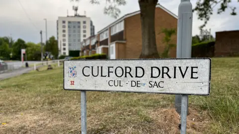 A street sign that says Culford Drive, cul de sac, is in the foreground. Behind the sign, slightly out of focus, is a patch of grass with a row of houses behind it, and a block of flats further in the background