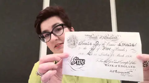 A woman in an indoor setting holds an old Bank of England five‑pound note toward the camera. The banknote shows ornate black calligraphy, decorative flourishes, and the text promising payment to the bearer. The note appears dated 1936, with a classical emblem and the printed signature of the Chief Cashier.