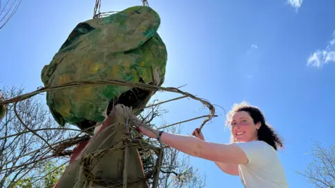 BBC/Andy Watson A large green puppet made of hessian and willow is being constructed by volunteers at the Upfront Puppet Theatre in Penrith. The picture depicts one of the volunteers standing with one of the large puppets in front of a bright blue sky. 
