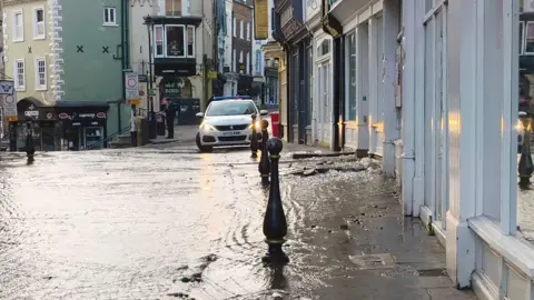 Claire Donlan Water flowing down Saddler Street in Durham. There are shop fronts on the right side of the narrow streets with restaurants and a police car in the distance.
