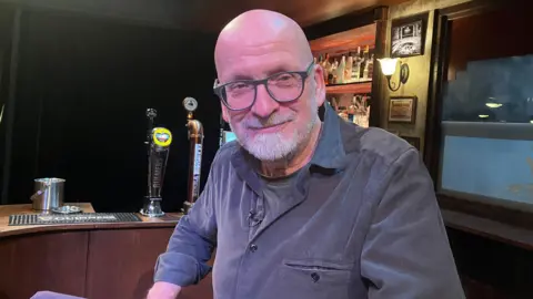 BBC A man with a bald head, glasses and a white beard sits in a bar on a theatre stage. He is wearing a grey t-shirt under a blue-grey shirt.