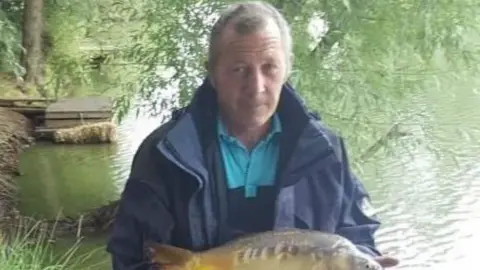 Family David Carpenter with a fish. He is on a bank and there is water and a tree behind him. He is wearing a coat, a blue polo neck and smiling. 