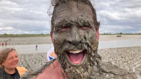Man's face covered in mud- he is smiling broadly and the estuary is behind him