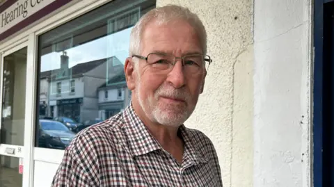 A man, wearing a flannel shirt and glasses. He is looking at the camera with a slight smile. He is stood in front of a shop. There is a cream wall and windows and a white door. The logo on the shop says "the New Hearing Centre"
