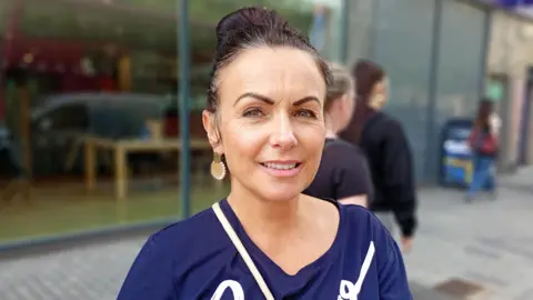 A woman, with brown hair tied up, wearing a navy top. She is looking at the camera. 