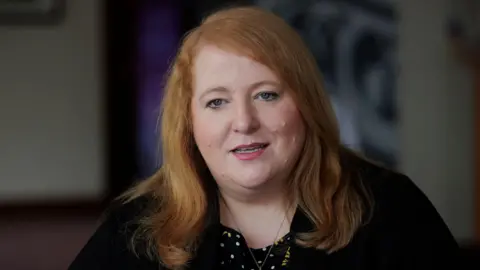 Naomi Long - a woman with shoulder-length, ginger hair is sat down. She is wearing a dark jacket, and a black-and-white polka dot tshirt with yellow foral pattern. The background of the room she is sitting in is blurred.