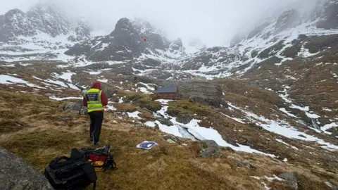 A drone pilot wearing a high visibility vest flies a drone in a mountain landscape with patches of snow.