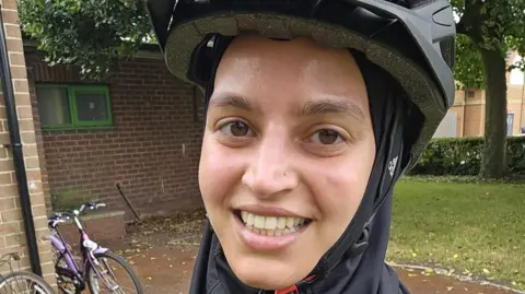 Zubeida Osman A woman with a nose stud smiles at the camera, wearing a black bike helmet and a black sport hijab underneath. She is standing in front of a brick building with a purple bicycle propped up against it