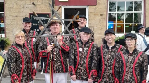 Grenoside Sword Dancers An image of seven sword dancers. They are looking at the camera and one of them is holding six swords in a star formation. They are smiling and wearing brightly patterned red and black outfits with gold detailing. They are standing outside a pub.