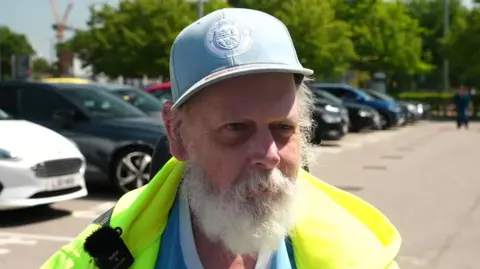 Brian Mitchell has white hair and white beard. He wears sky blue branded Manchester City baseball cap and yellow high-visibility anorak. There are cars behind him. He is the focus on the photo.