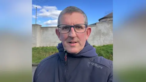 Gareth Barker Gareth Barker has short salt and pepper coloured hair and black framed glasses.  He is wearing a zipped up navy coat