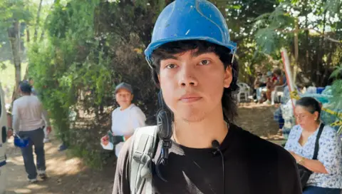 BBC News Mundo Alejandro Mahecha, he's a young person wearing a blue hard hat and a black t-shirt - he has medium length hair and has a serious expression 
