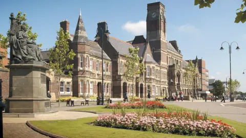 St Helens Borough Council A flowerbed on a verge lit up by the sun give ways to a view of the historical St Helens Borough Council building, which features spires and a clock tower. 