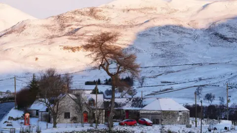 PA Media A couple of homes in Glenshee, with two red cars sitting in front of them. A red phone box is outside and hills are behind the homes. Snow covers the grounds. 