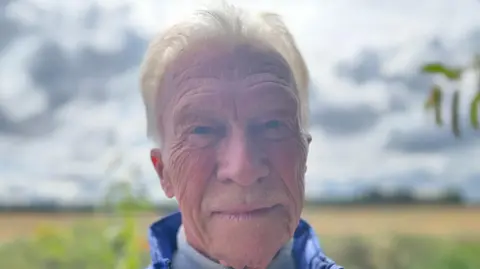 John Jackson, who has light grey hair and rosy cheeks, looks towards the camera with a serious expression on his face. He is wearing a dark blue gilet.