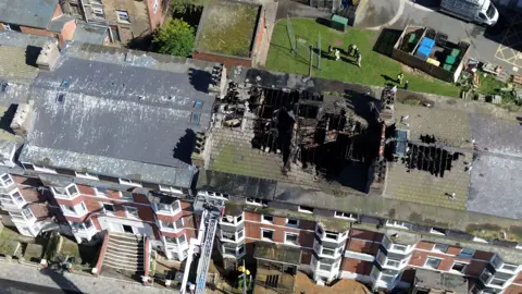 An aerial view of a row of five-storey brick-terraced buildings following a fire. The roof of the middle property shows extensive damage, with large sections burned through and collapsed, exposing blackened beams and interior spaces. Adjacent roof on right appears slightly damaged with black staining. A fire service aerial ladder is extended from street level up to the front of the middle building. Behind the properties, small gardens and yards are visible, with a few people standing on a grassy area nearby. The scene is in daylight.
