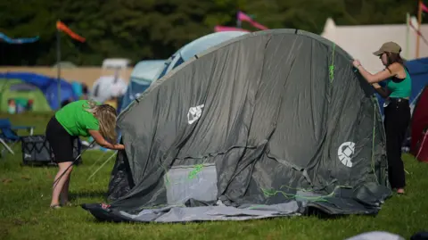 PA Two women set up a large grey tent at Glastonbury Festival. 