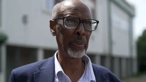 Eid Ali Ahmed stands outside the conference venue. He's smartly dressed in a navy suit, and black rimmed glasses. He has a neatly trimmed grey beard and cropped white hair. 