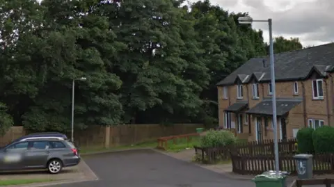 A residential street with cars parked on one side and a number of terraced homes on the other.