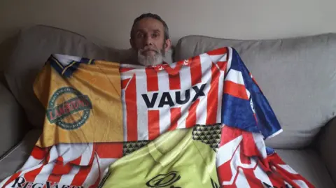 Handout Paul Fairless sits on a grey sofa and is holding up a blanket. The blanket is made up of various Sunderland football shirts stitched together. 
