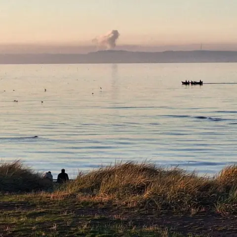 Jane Patmore Two people sit on the bank of a river. A small boat is sailing in the background, which includes a large cloud
