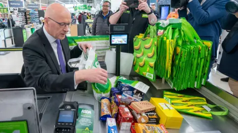 PA Media John Swinney is sitting at an Asda checkout where he is wearing a dark suit, white shirt and purple tie. He is bagging a pile of shopping as photographers and camera people capture the image.