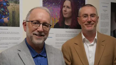 Chief Executive Officer at Nottingham University Hospitals, Anthony May stands alongside photography Stephen Iliffe standing in front of the exhibiton. Both men are wearing glasses, Anthony on the left is in a grey suit blazer while Stephen has a brown suit jacket on. 