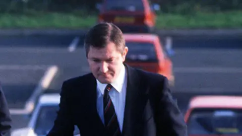 Pacemaker Pat Finucane is walking away from a car. He is wearing a suit and tie and has short brown hair.