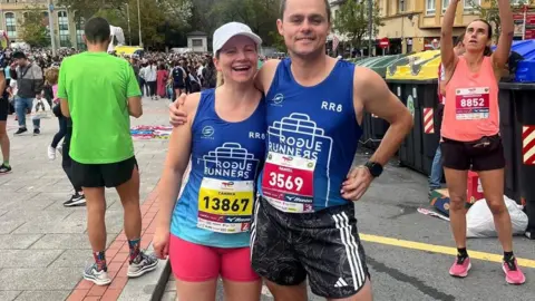 Family handout A woman and a man in running gear. There is a runner behind them taking a photo and there is a crowd behind them. 