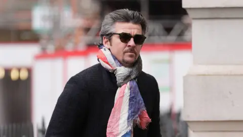 Joey Barton arriving at Westminster Magistrates' Court on 24 January. Mr Barton, who has grey hair and a goatee, is wearing sunglasses, a red white and blue scarf and a dark tweed coat