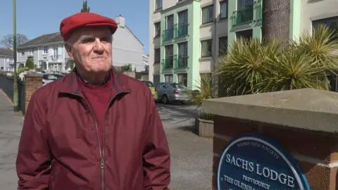 An elderly man in a burgundy jacket and a red flat cap, stood alongside a blue plaque saying "Sachs Lodge"