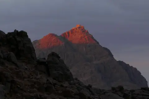 Ben Hoffler The peak of Mount Sinai at dusk in 2024. The light catches the top of the rocky mountain, which is standing higher than another mountain in the foreground
