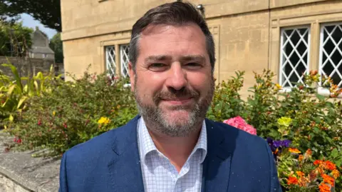 Kevin Guy. He has short dark hair and a beard. He is wearing a checked shirt and a navy blue jacket. He is looking at the camera and smiling.